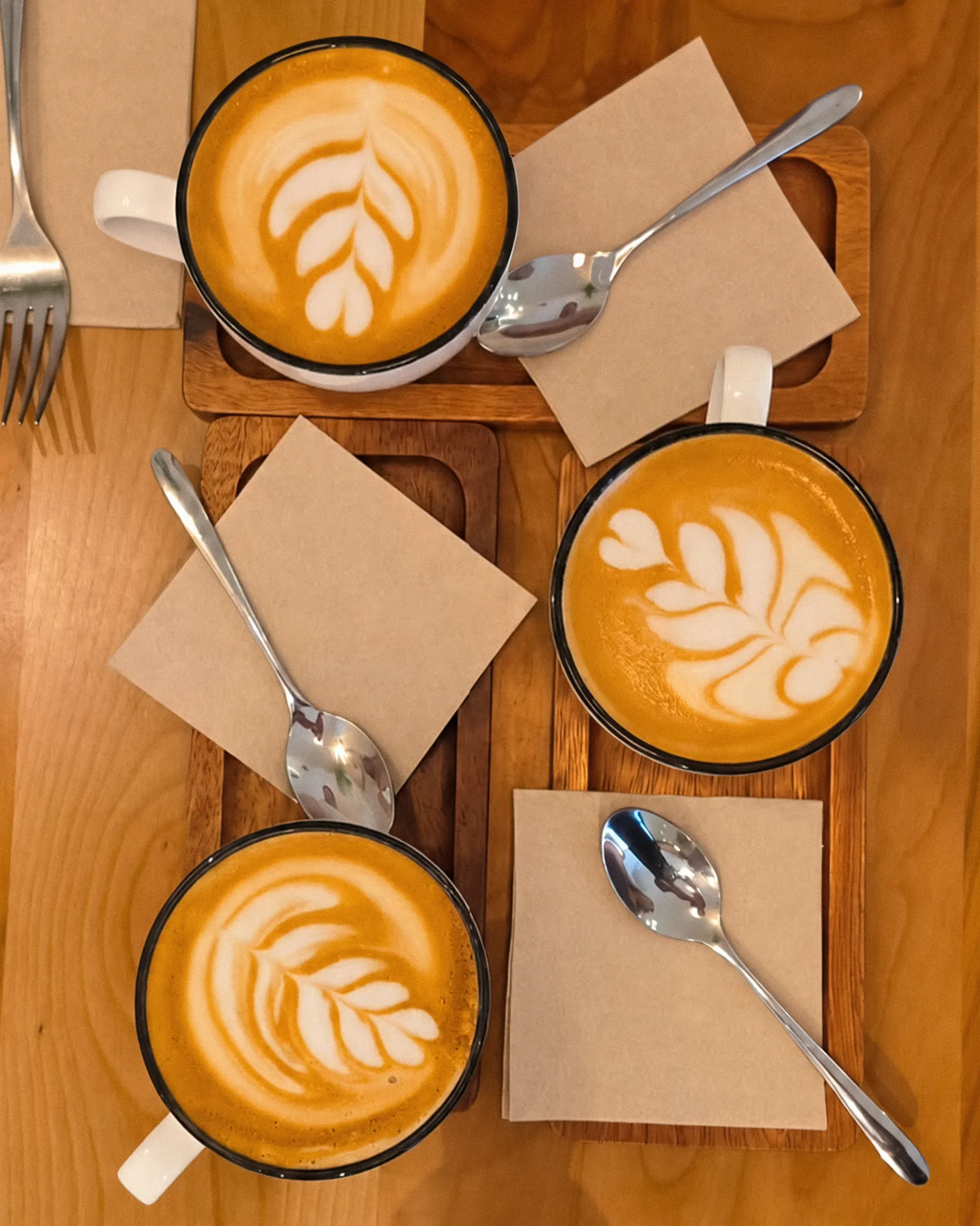 Tres cafés con leche con latte art en forma de hoja, sobre bandejas de madera, vistos desde arriba