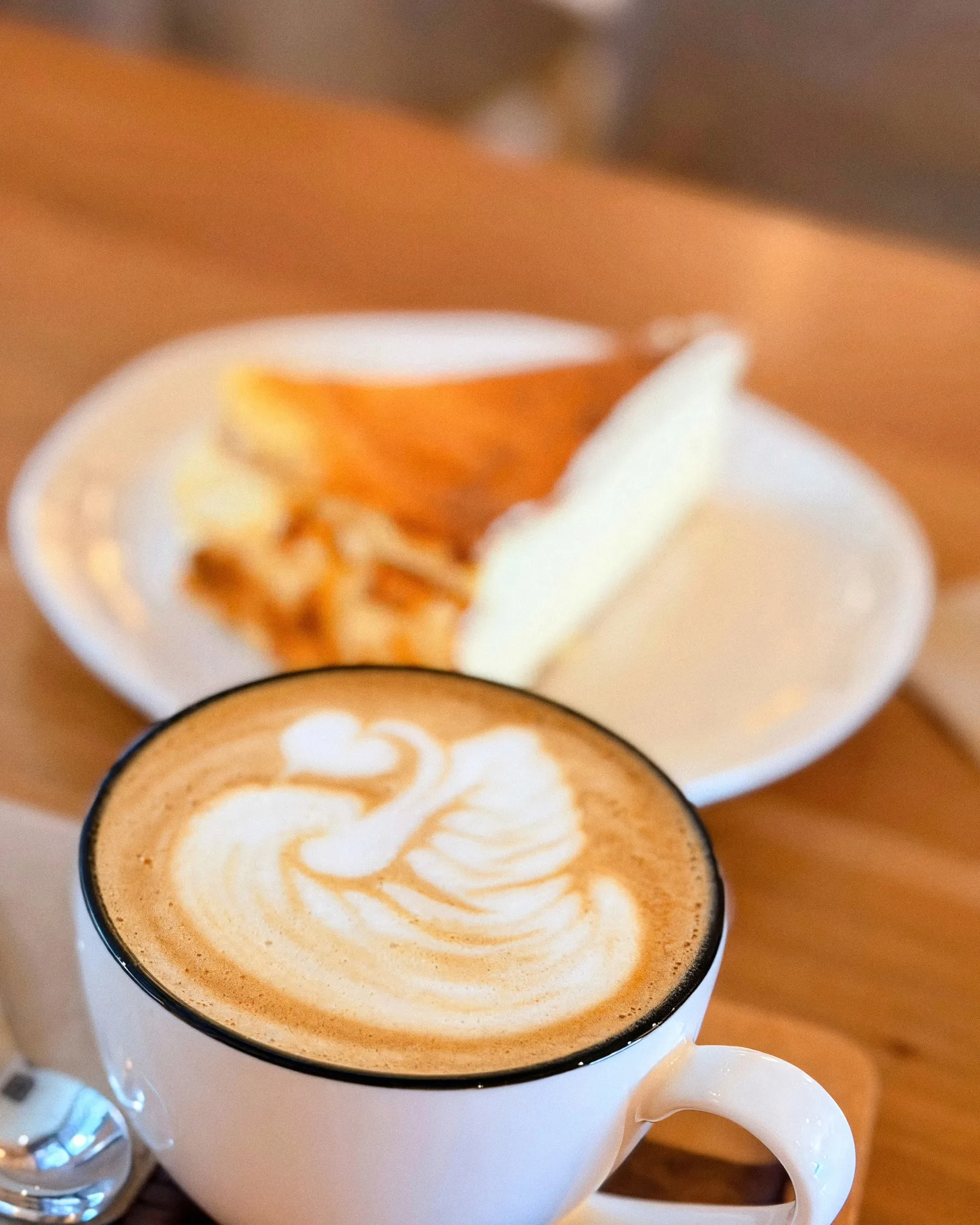 Cappuccino con un cisne de latte art y, detrás, una porción de cheesecake