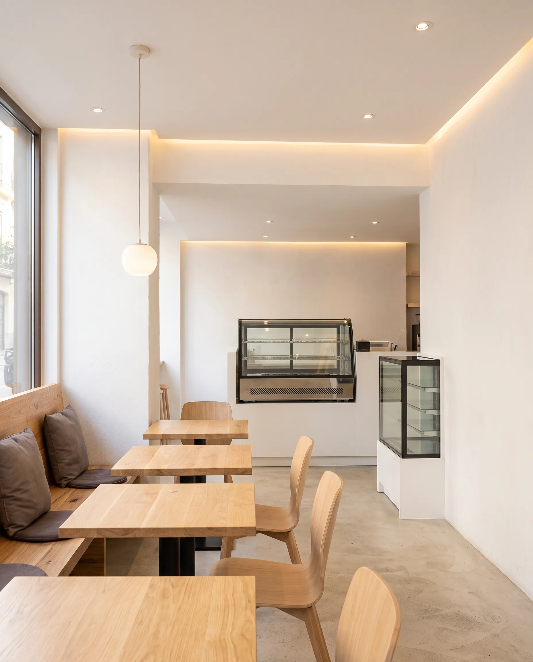 Sala luminosa con mesas y sillas de madera clara, banco y vitrina al fondo